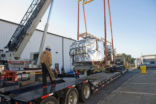 A contractor prepares the pallet holding Helios 1 to be lifted by a 70-ton crane so Helios can be loaded onto a flatbed tractor-trailer and begin its trip to Singapore. The loading procedure took place Dec. 21. Helios 1 includes the 57,000 pound storage ring and three transportainers of support parts and equipment.