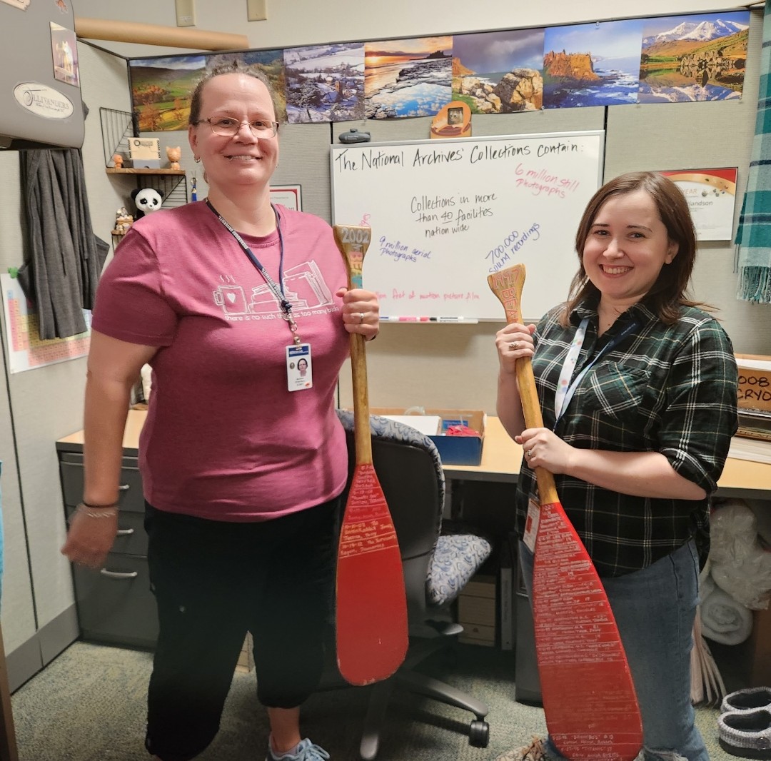 Information Resources staff Melissa Erlandson (left) and intern Kate McDannold display oars which contain names of the winning Beams teams including the school, team name and list of students, and their score. The BEAMS game consisted of designing a boat and placing marbles in it while floating in water.  The team whose boat held the most marbles won.  The oars were created and updated by Dave Kausch (Fire Prevention Manager) in 1997, 1998, and 2002.