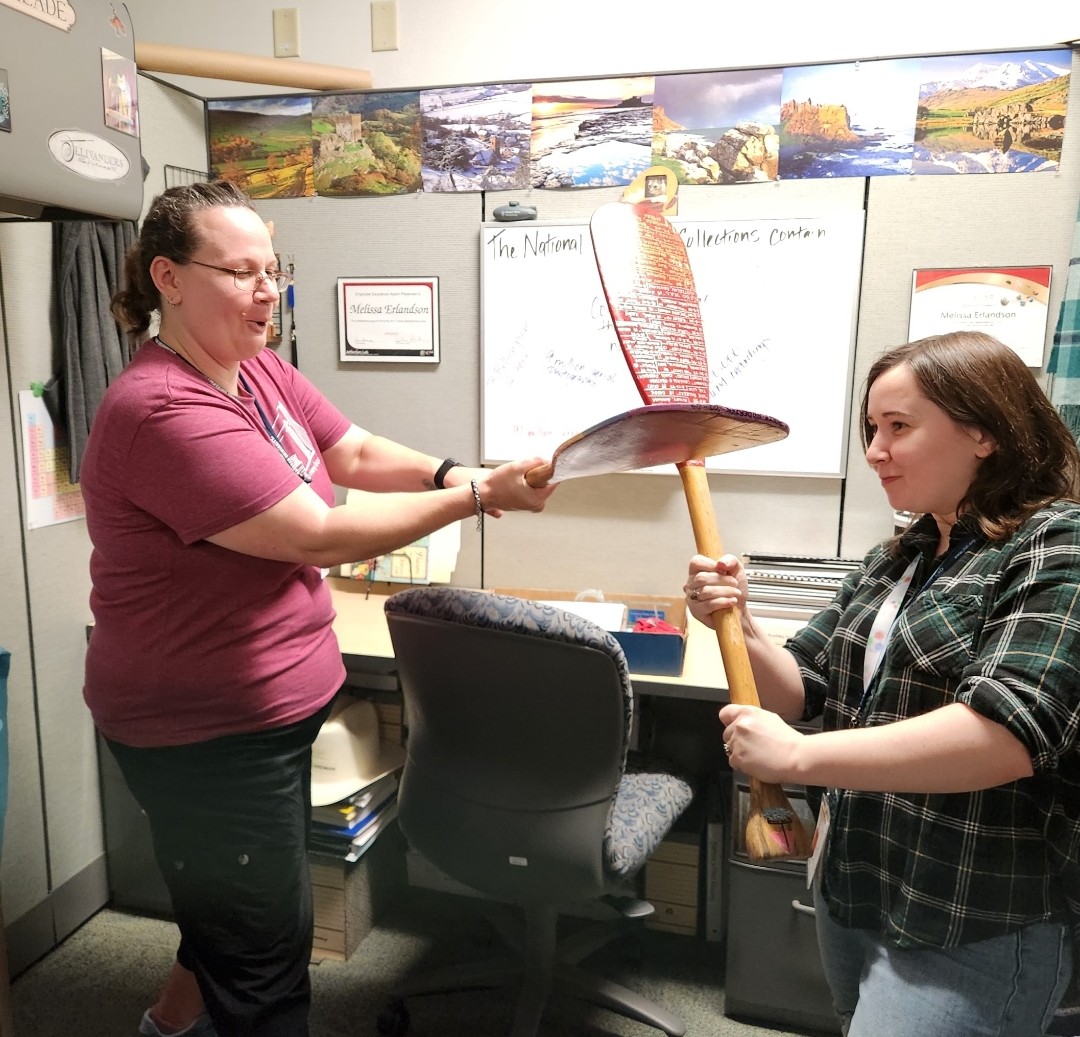Information Resources staff Melissa Erlandson (left) and intern Kate McDannold display oars which contain names of the winning Beams teams including the school, team name and list of students, and their score. The BEAMS game consisted of designing a boat and placing marbles in it while floating in water.  The team whose boat held the most marbles won.  The oars were created and updated by Dave Kausch (Fire Prevention Manager) in 1997, 1998, and 2002.
