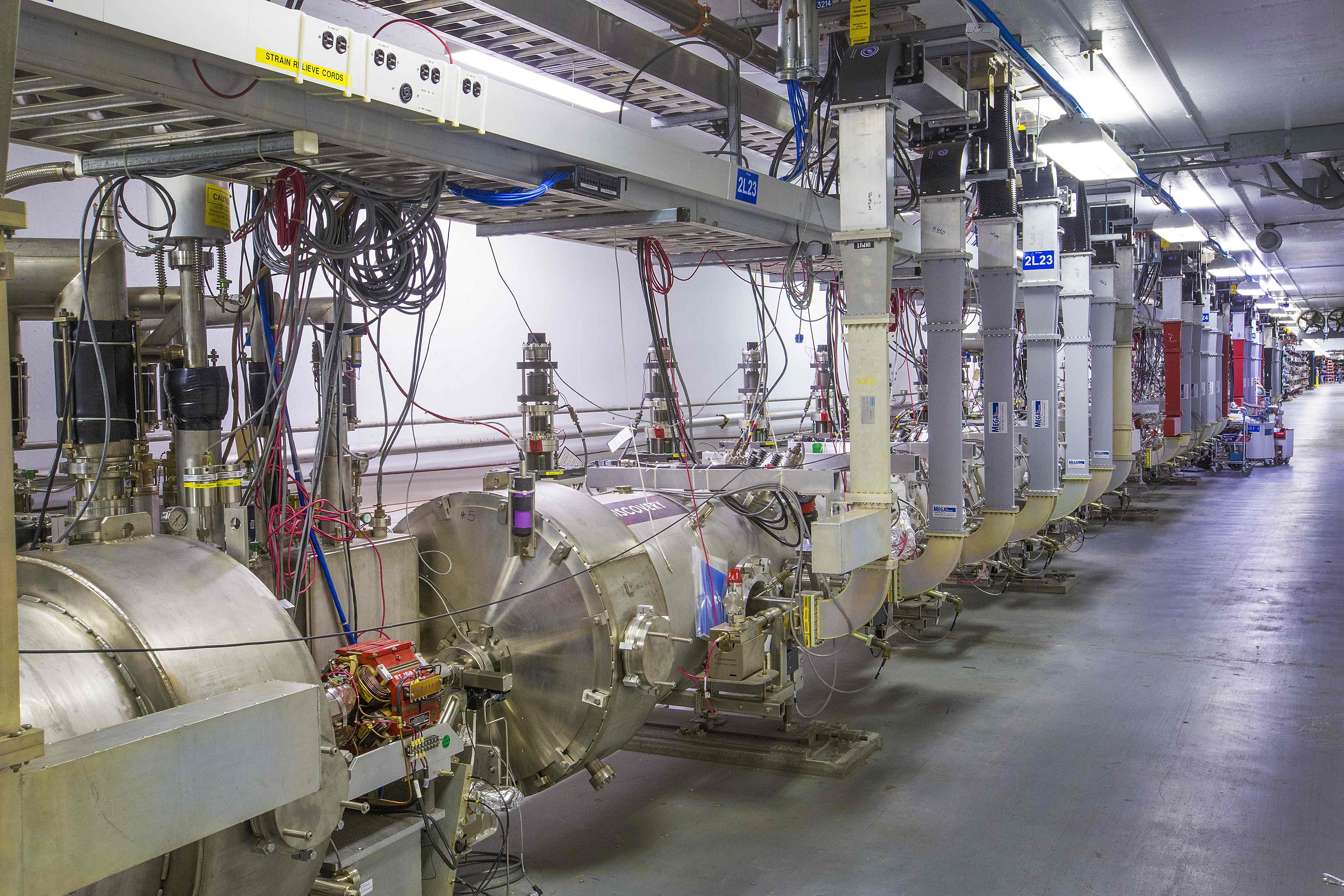 Photo of CEBAF accelerator, featuring a narrow concrete tunnel with acceleration modules that are contained in stainless steel sleeves that are roughly three feet in diameter and thirty feet long.