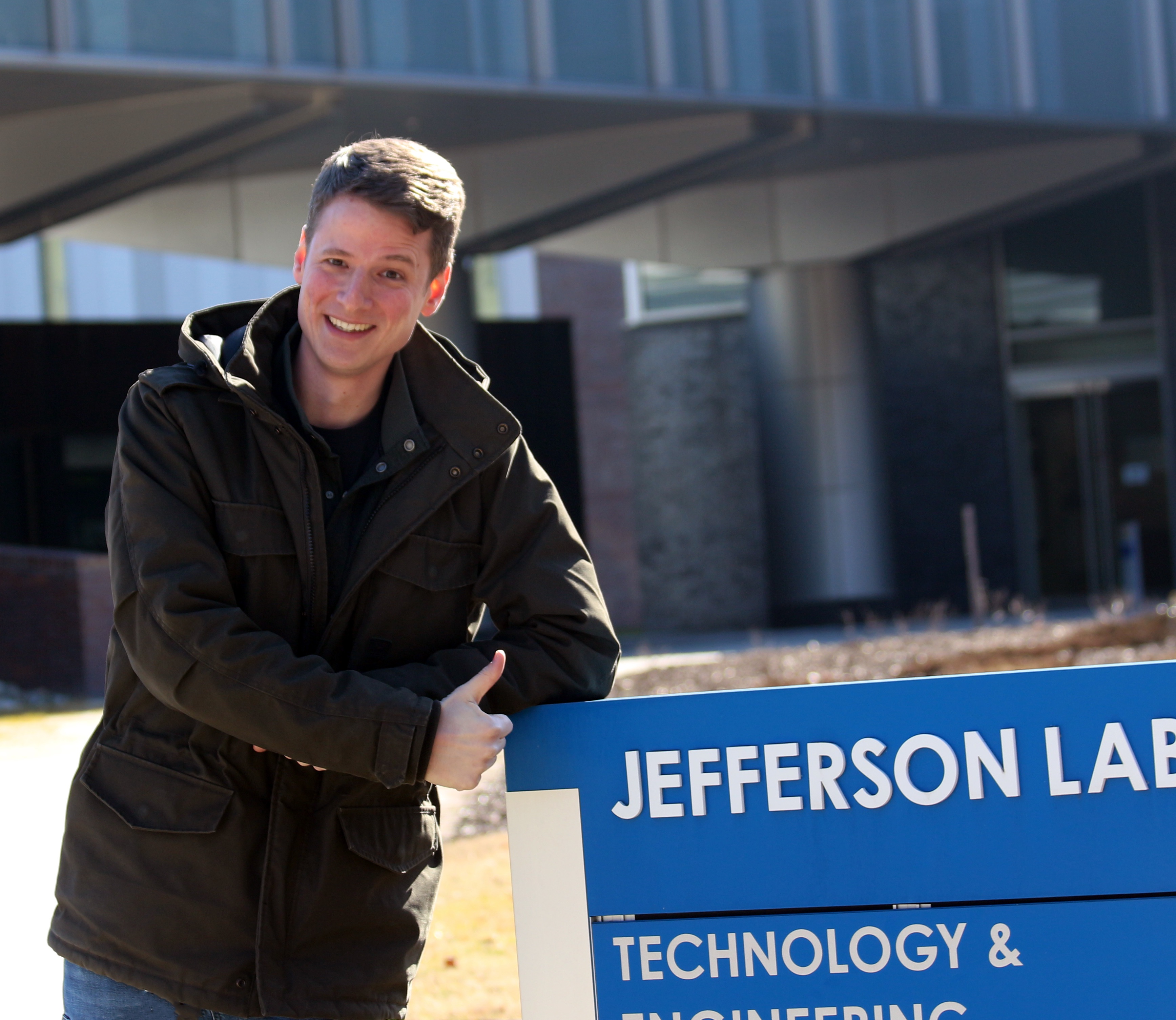John Vennekate next to the blue building sign for the Jefferson Lab Technology and Engineering Development building
