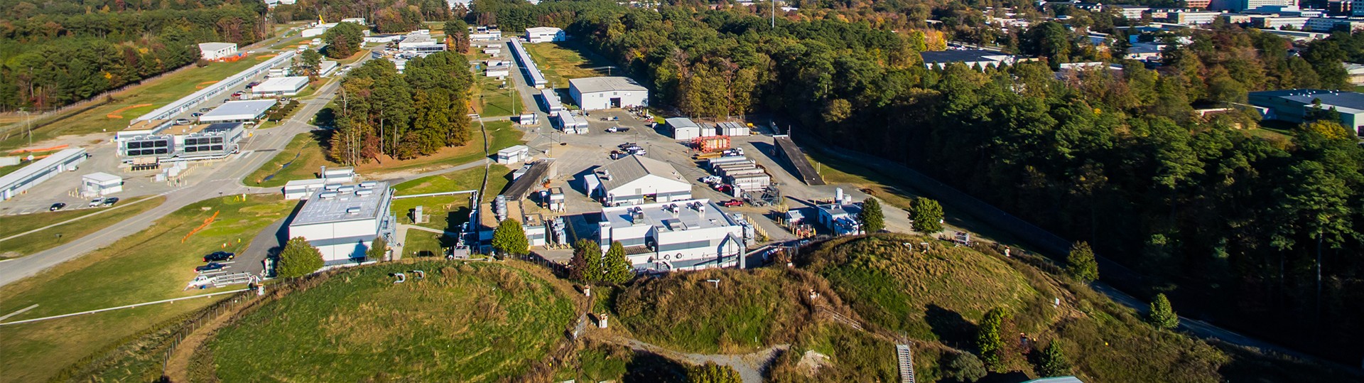 Aerial photo of the Continuous Electron Beam Accelerator Facility, showing the above-ground accelerator service buildings, which outline the accelerator's racetrack shape. The three round domes of the Experimental Halls A, B and C are in the foreground, and the Experimental Hall D complex is in the top left corner of the photo.