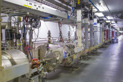 Photo of CEBAF accelerator, featuring a narrow concrete tunnel with acceleration modules that are contained in stainless steel sleeves that are roughly three feet in diameter and thirty feet long.
