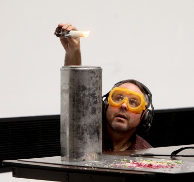Science Steve prepares to dip a short, white candle that has been lit into the top of a narrow cylindrical dewar that contains a small amount of liquid nitrogen. He is wearing safety goggles and ear protection headphones.