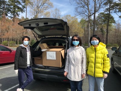 Three women in masks show boxes of supplies donated in car trunk